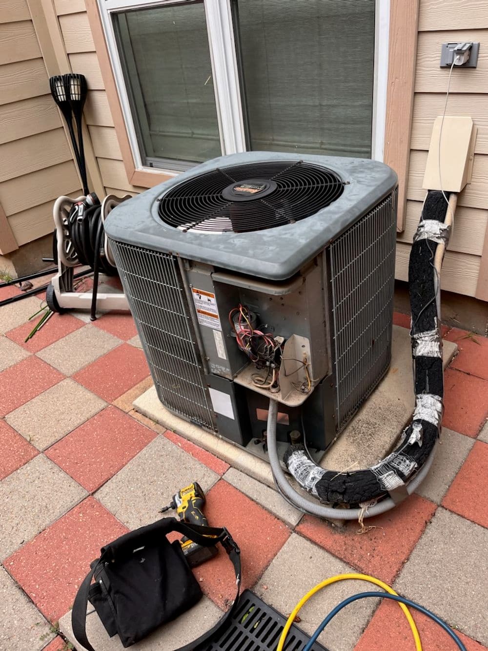 Air conditioning unit outside a house with tools and equipment nearby on a stone patio.